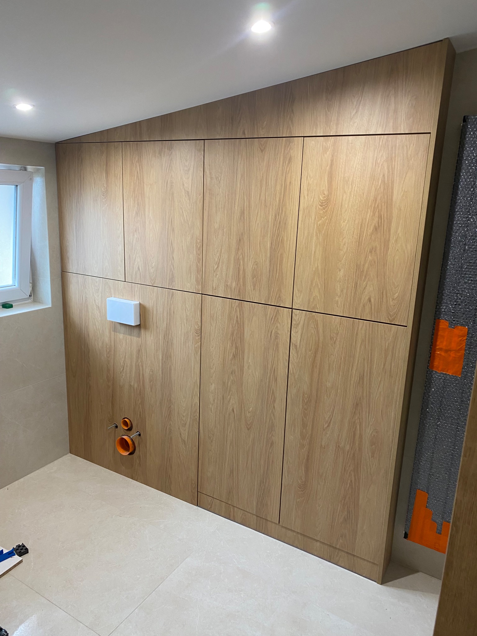 Drewniana zabudowa ściany w łazience z ukrytą spłuczką i instalacją sanitarną. Widoczne płytki podłogowe i fragment okna. Jasne oświetlenie.