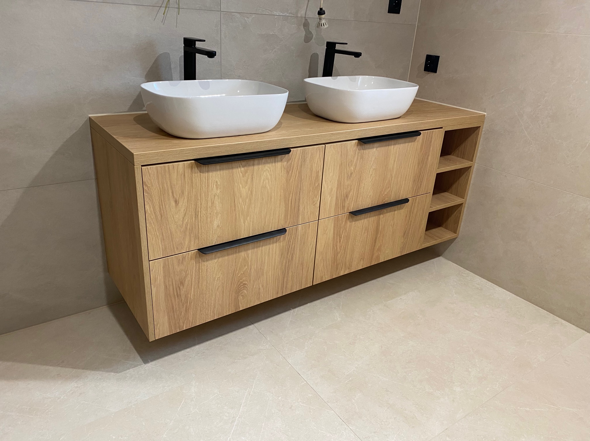 Łazienkowa szafka z drewnianymi frontami, czarnymi uchwytami i dwiema białymi umywalkami nablatowymi. Dodatkowe otwarte półki z boku szafki. Beżowe płytki na ścianie i podłodze.