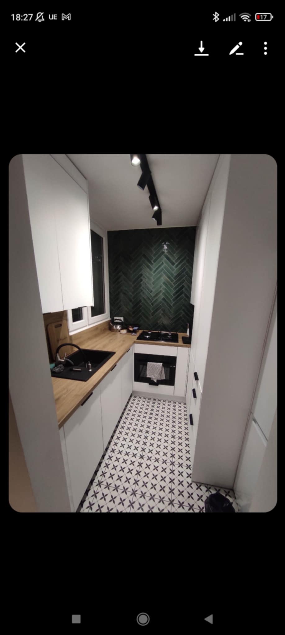 Wąska kuchnia z białymi szafkami i drewnianym blatem. Ściana z zielonych płytek ułożonych w jodełkę. Czarno-biała podłoga w geometryczny wzór, oświetlenie szynowe.