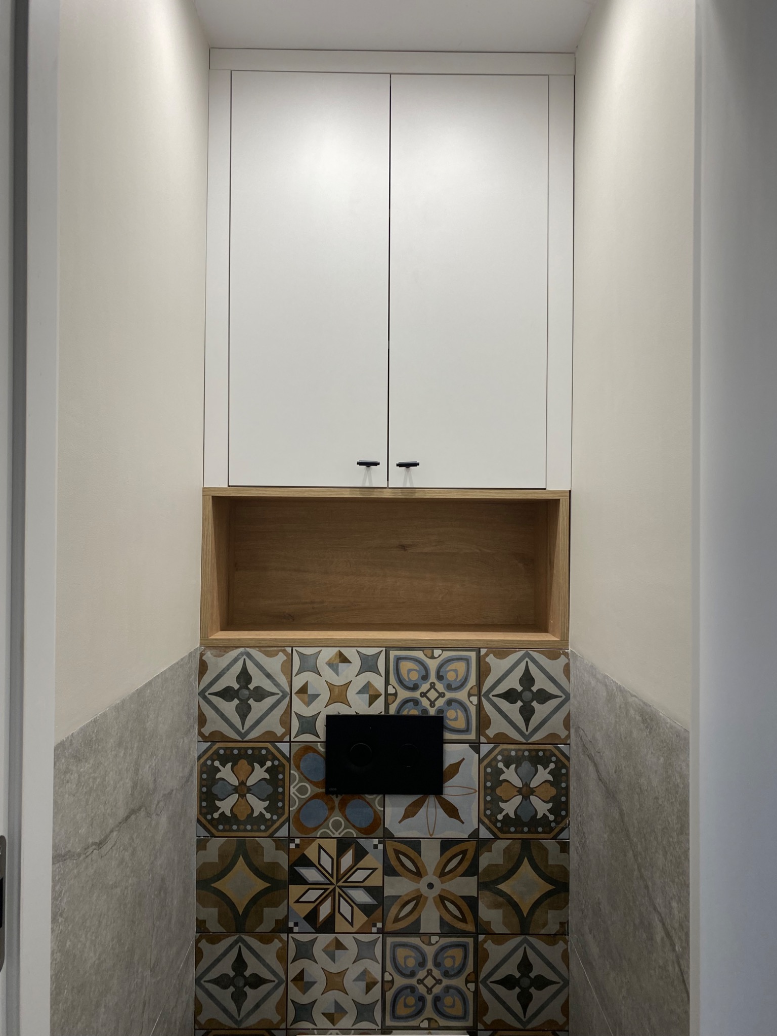 Biała szafka wisząca z drewnianą półką nad wzorzystymi płytkami w łazience, czarny przycisk spłuczki, neutralne ściany, minimalistyczny design.