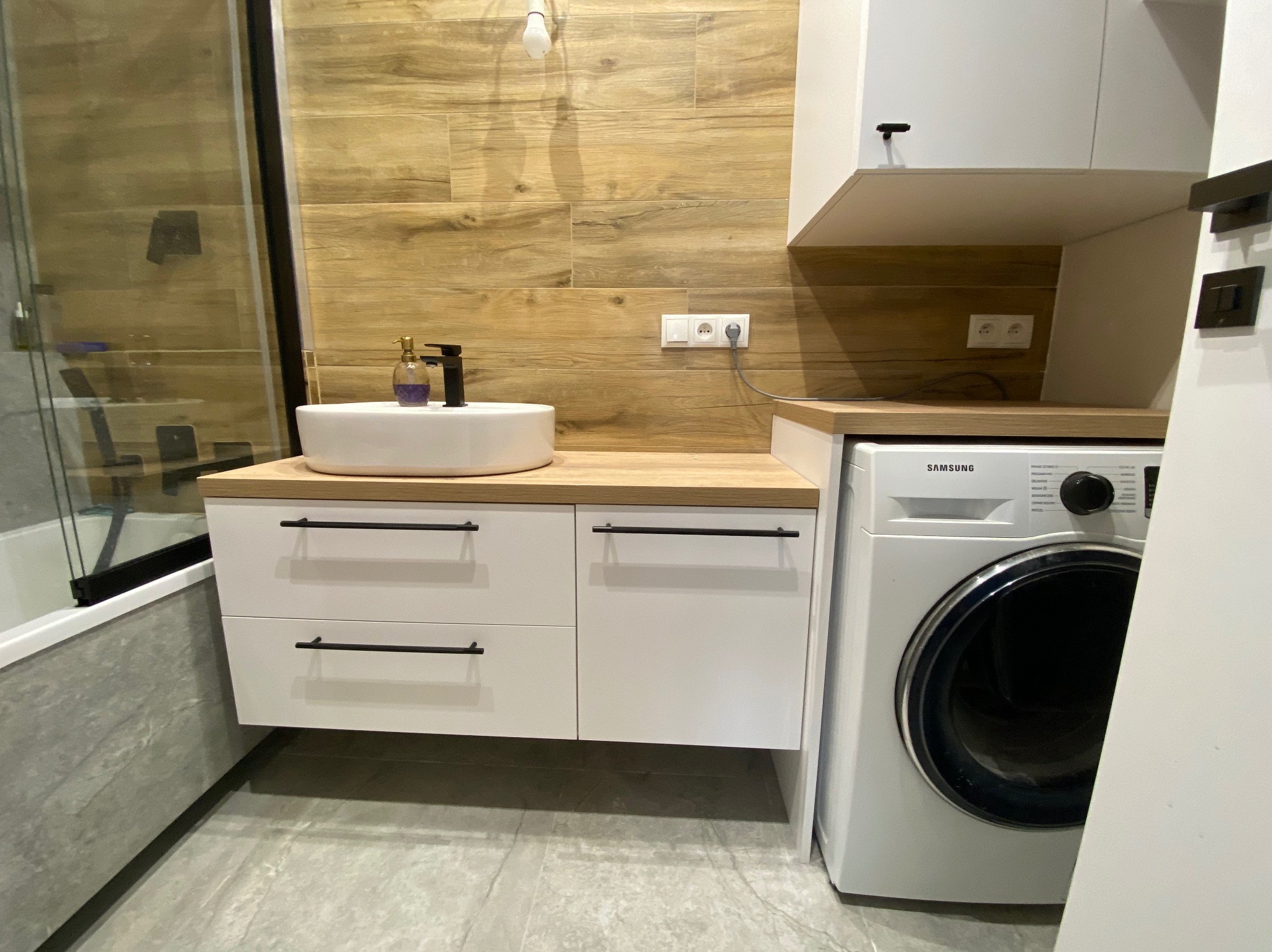 Nowoczesna łazienka z drewnopodobnymi płytkami, białą szafką pod umywalką z czarnymi uchwytami i pralką Samsung. Minimalistyczny design, ciepłe oświetlenie.