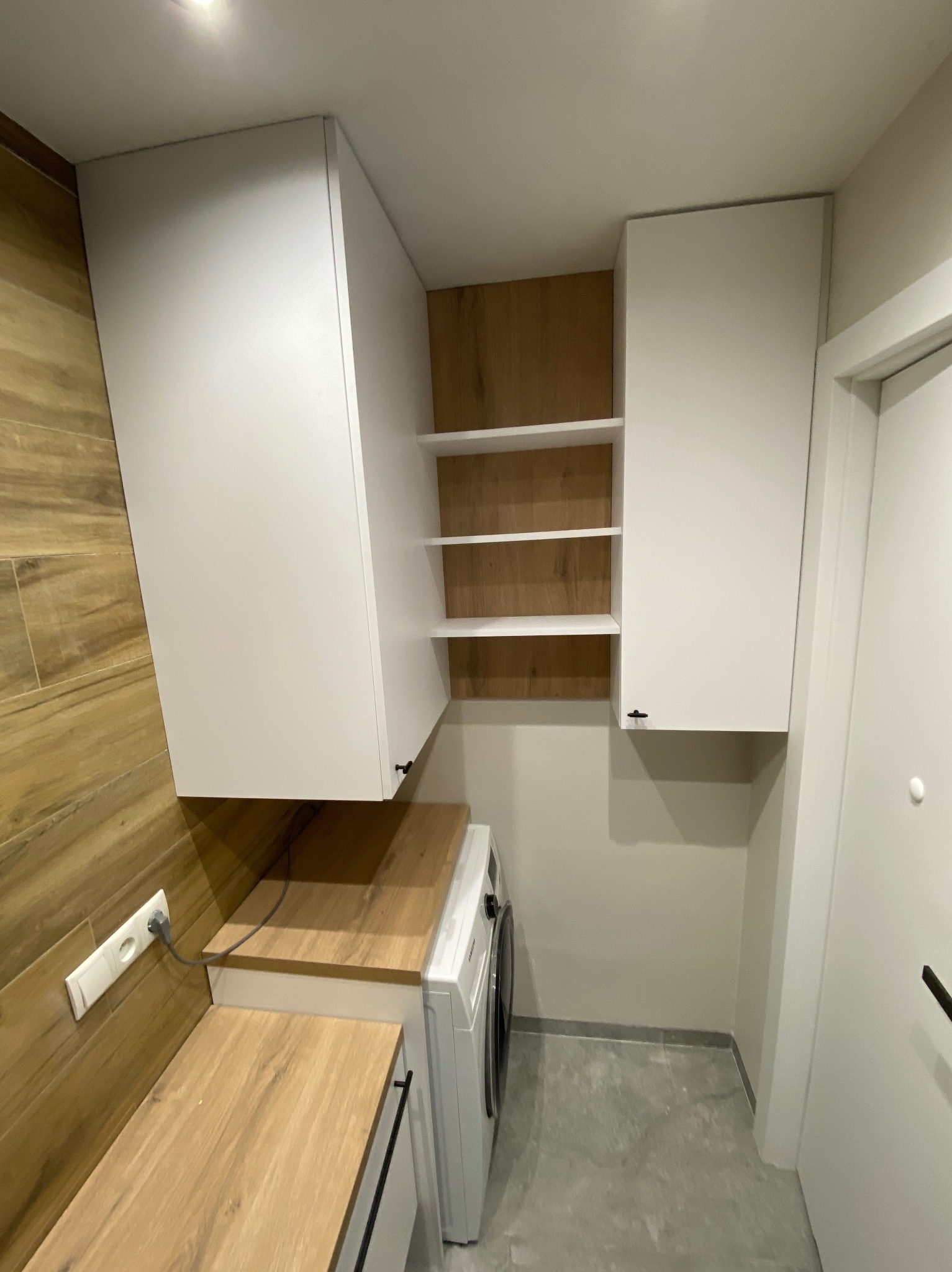 Białe szafki wiszące z drewnianymi blatami i półkami nad pralką w małej łazience. Minimalistyczny design, jasne oświetlenie, płytki imitujące drewno na ścianie.