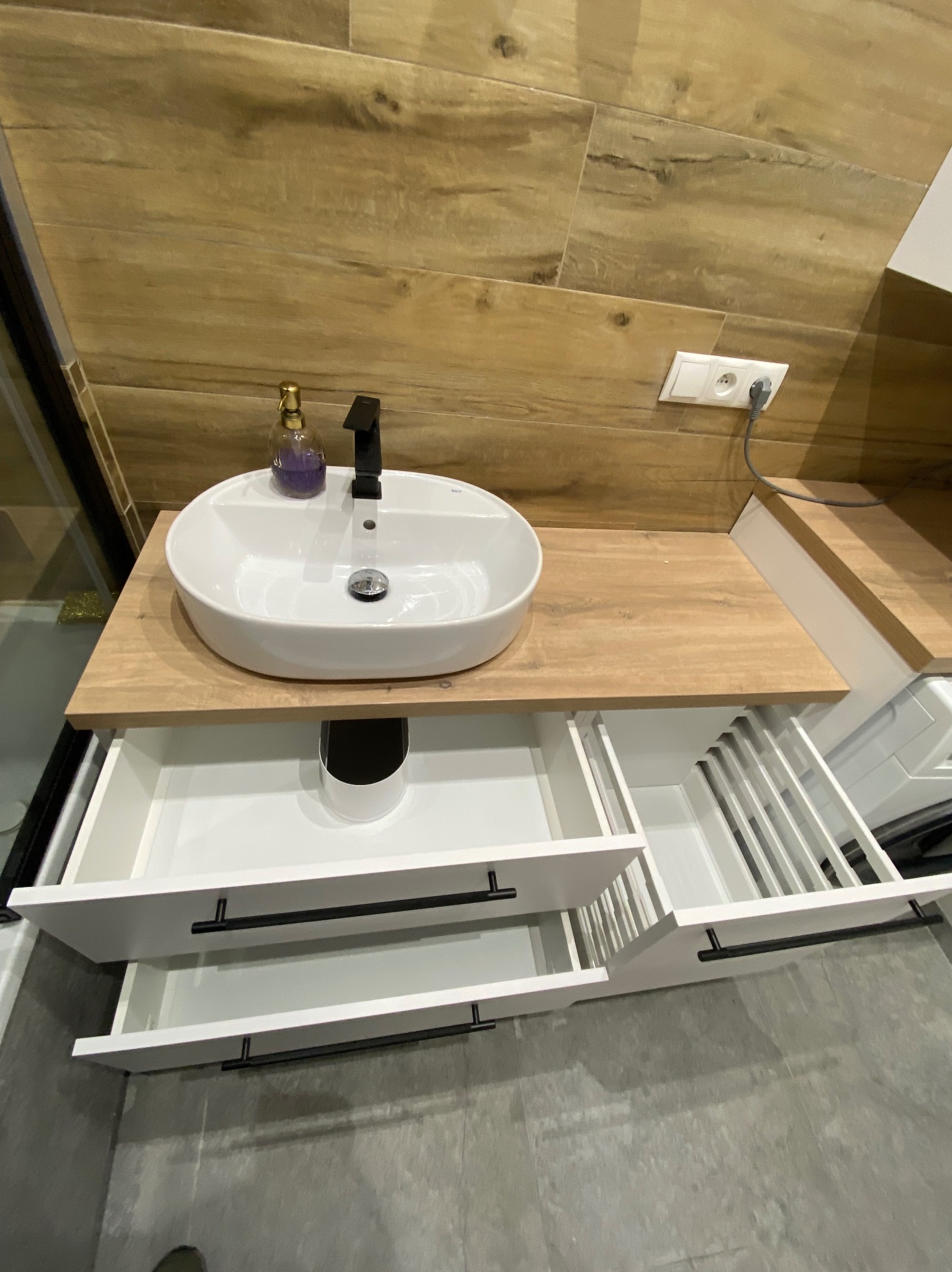 Nowoczesna łazienka: biała umywalka nablatowa na drewnianym blacie z otwartymi szufladami z czarnymi uchwytami. Na blacie fioletowe mydło w płynie.