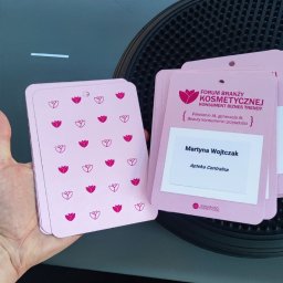 Makowski Design - Różowe identyfikatory z logo i napisem 'Forum Branży Kosmetycznej' leżące na czarnym tle, w dłoni.