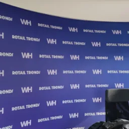 Niebieska ścianka reklamowa z powtarzającym się białym napisem 'RETAIL TRENDS WH', widoczny fragment krzesła biurowego. Tło dla konferencji lub prezentacji.