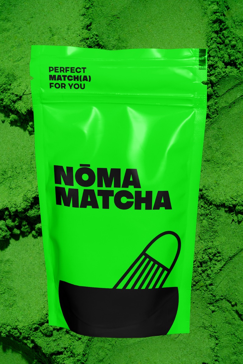 Jasnozielone opakowanie Nōma Matcha na tle sproszkowanej zielonej herbaty. Minimalistyczny design z czarną grafiką miseczki i ubijaczki.