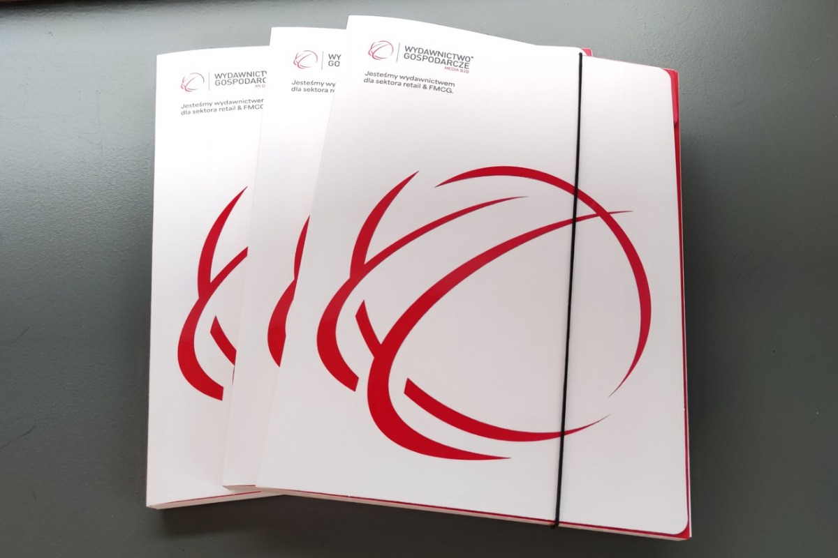 Trzy białe teczki z czerwonym logo i napisem 'Wydawnictwo Gospodarcze', spięte czarną gumką, leżą na szarym tle. Teczki lekko przesunięte względem siebie.