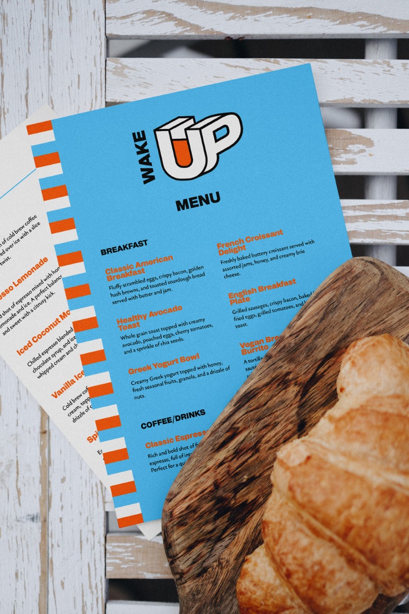 Menu restauracji 'Wake Up' z ofertą śniadaniową i kawową, leżące na drewnianej desce z croissantem, umieszczone na tle białego, drewnianego stołu.
