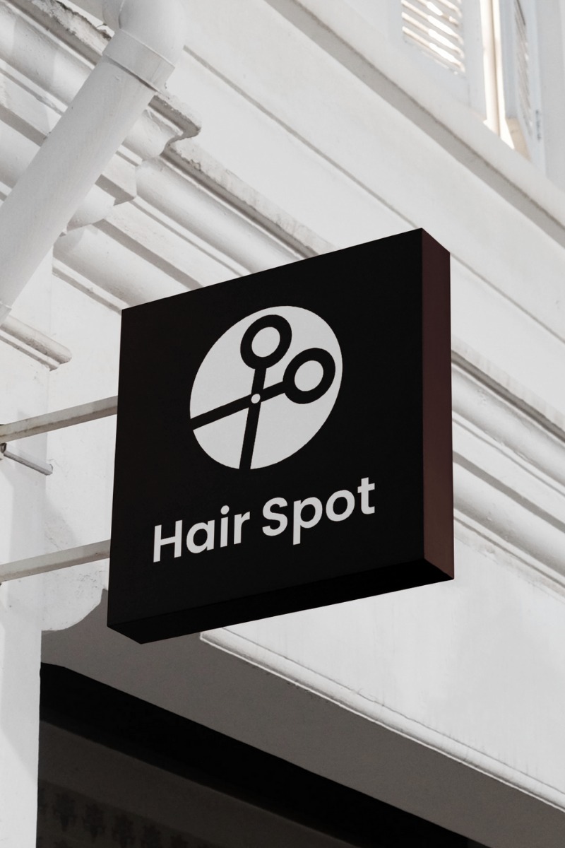 Czarny szyld salonu fryzjerskiego 'Hair Spot' z białym logo nożyczek w okręgu, zamontowany na białej fasadzie budynku. Minimalistyczny design, kątowe ujęcie.