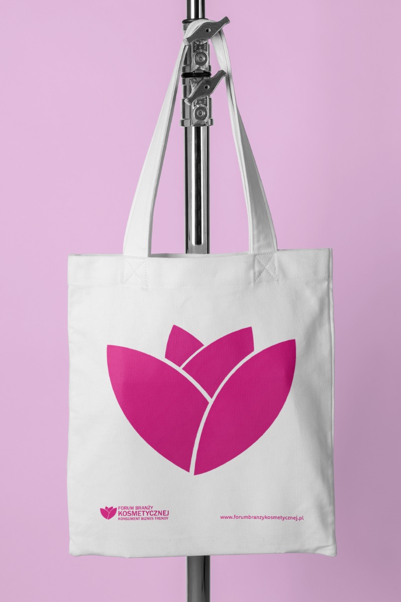 Biała torba bawełniana z różowym logo i adresem strony internetowej, zawieszona na metalowym stojaku na tle jasnoróżowego tła.