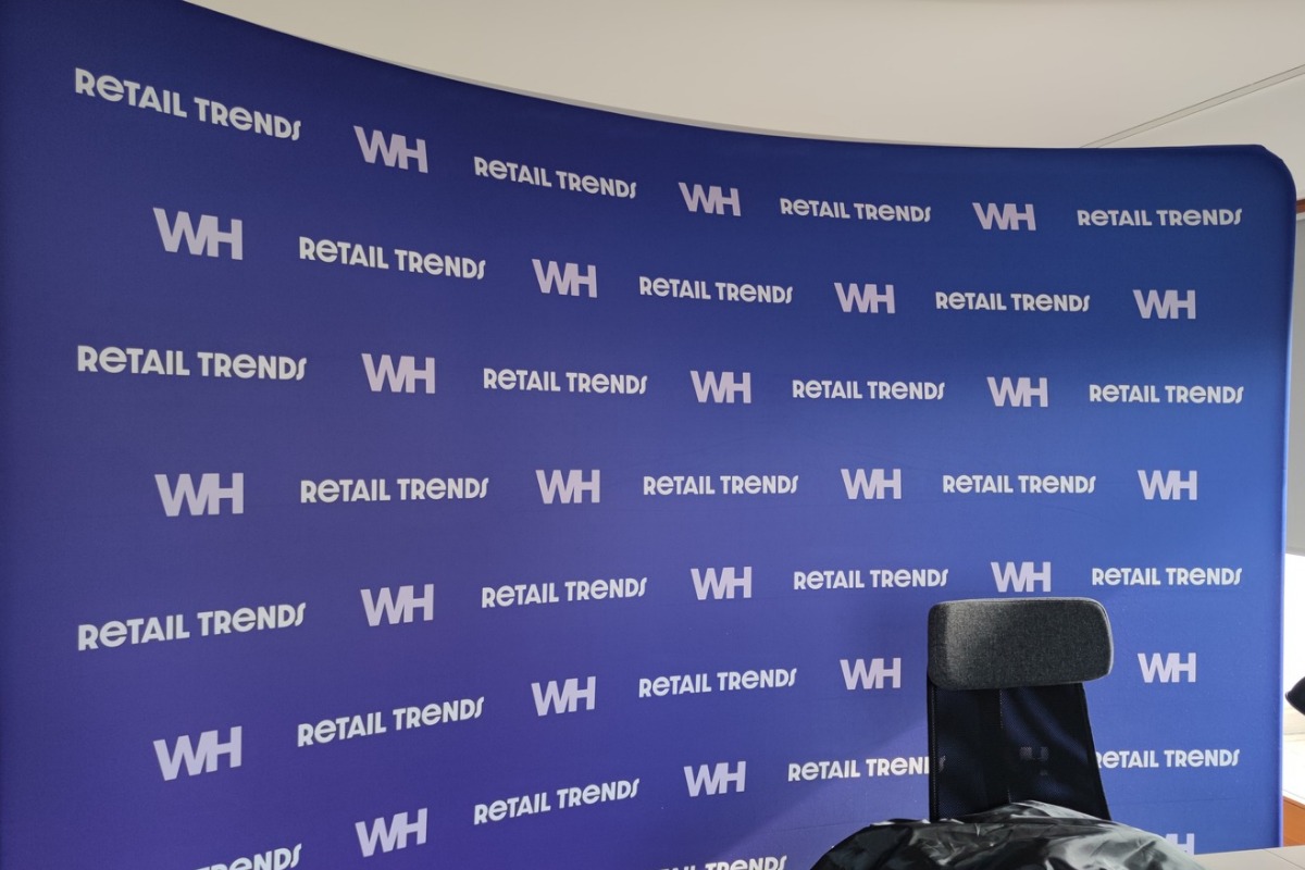 Niebieska ścianka reklamowa z powtarzającym się białym napisem 'RETAIL TRENDS WH', widoczny fragment krzesła biurowego. Tło dla konferencji lub prezentacji.