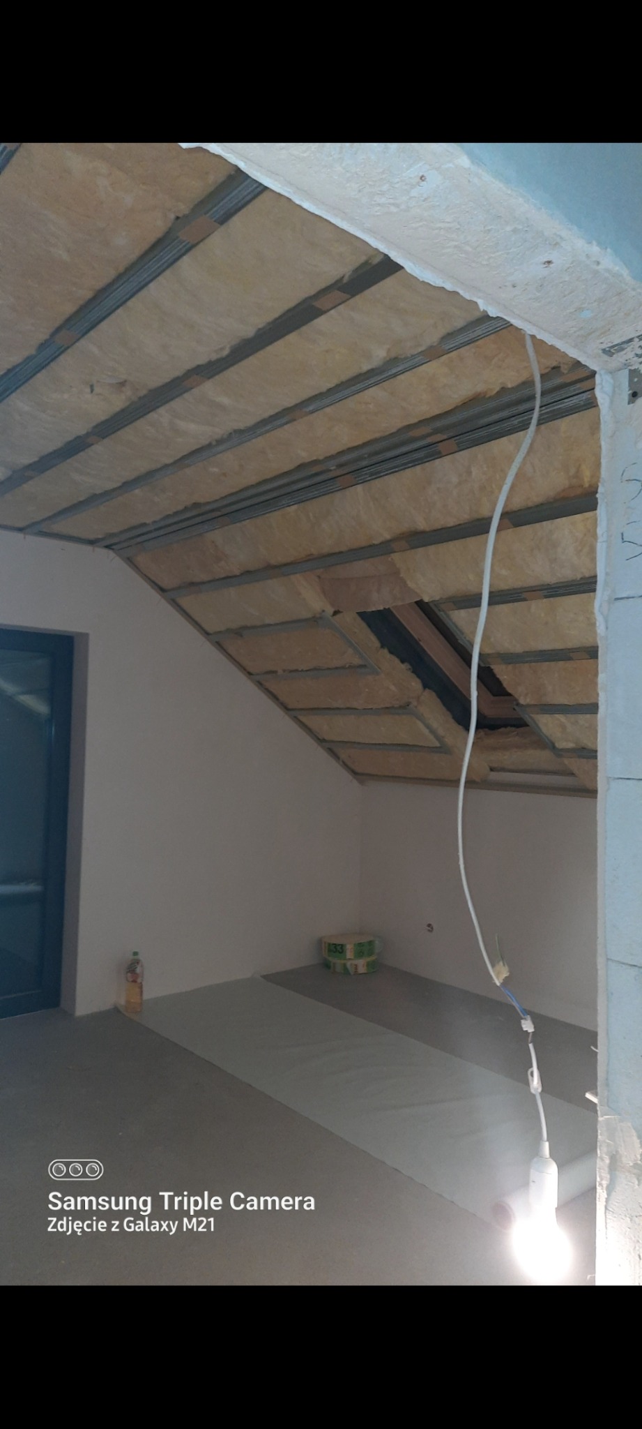 Izolacja poddasza wełną mineralną na metalowej konstrukcji. Widoczny fragment okna i podłoga. Oświetlenie tymczasowe.