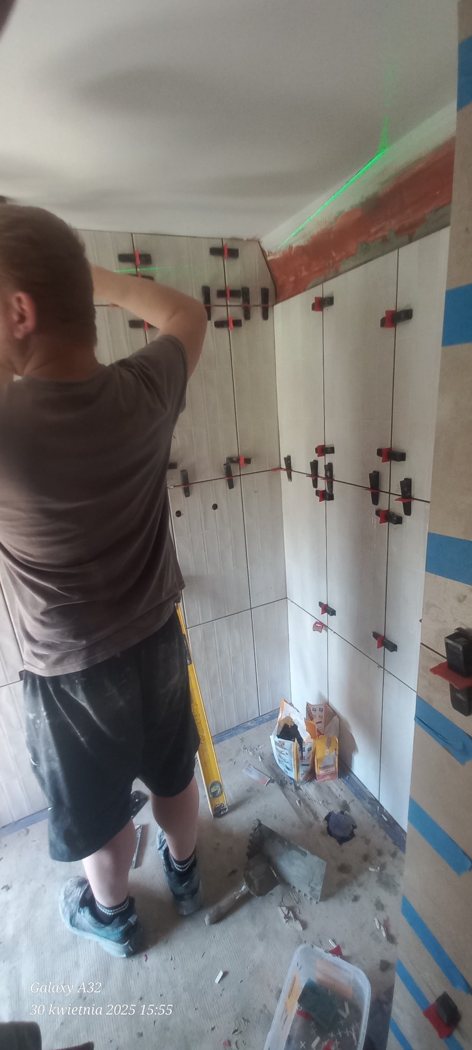 Układanie białych płytek w rogu pomieszczenia z użyciem lasera i krzyżyków dystansowych. Widoczne narzędzia glazurnicze i worki z zaprawą.
