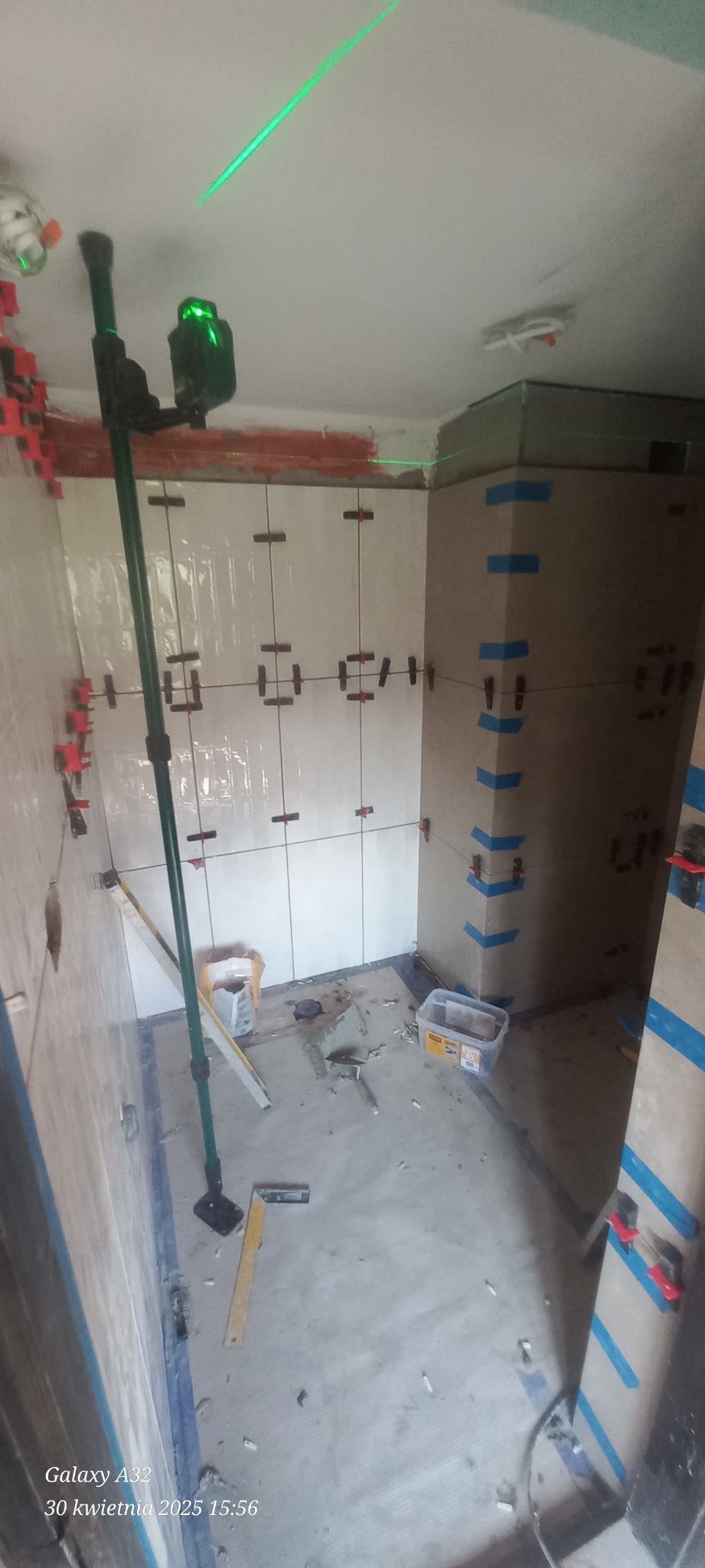 Układanie białych płytek w łazience, widoczny laser krzyżowy, poziomica i system poziomowania płytek. Prace glazurnicze w toku, surowy stan podłogi.
