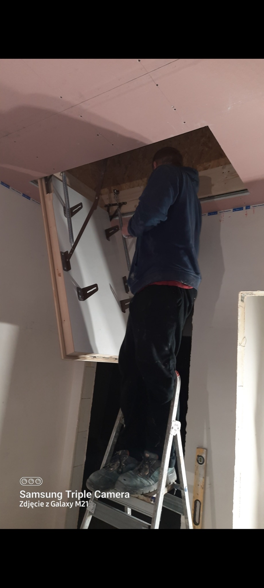 Montaż schodów strychowych: mężczyzna na drabinie instaluje składane schody w suficie, widoczne elementy mechaniczne i sufit z płyt gipsowo-kartonowych.