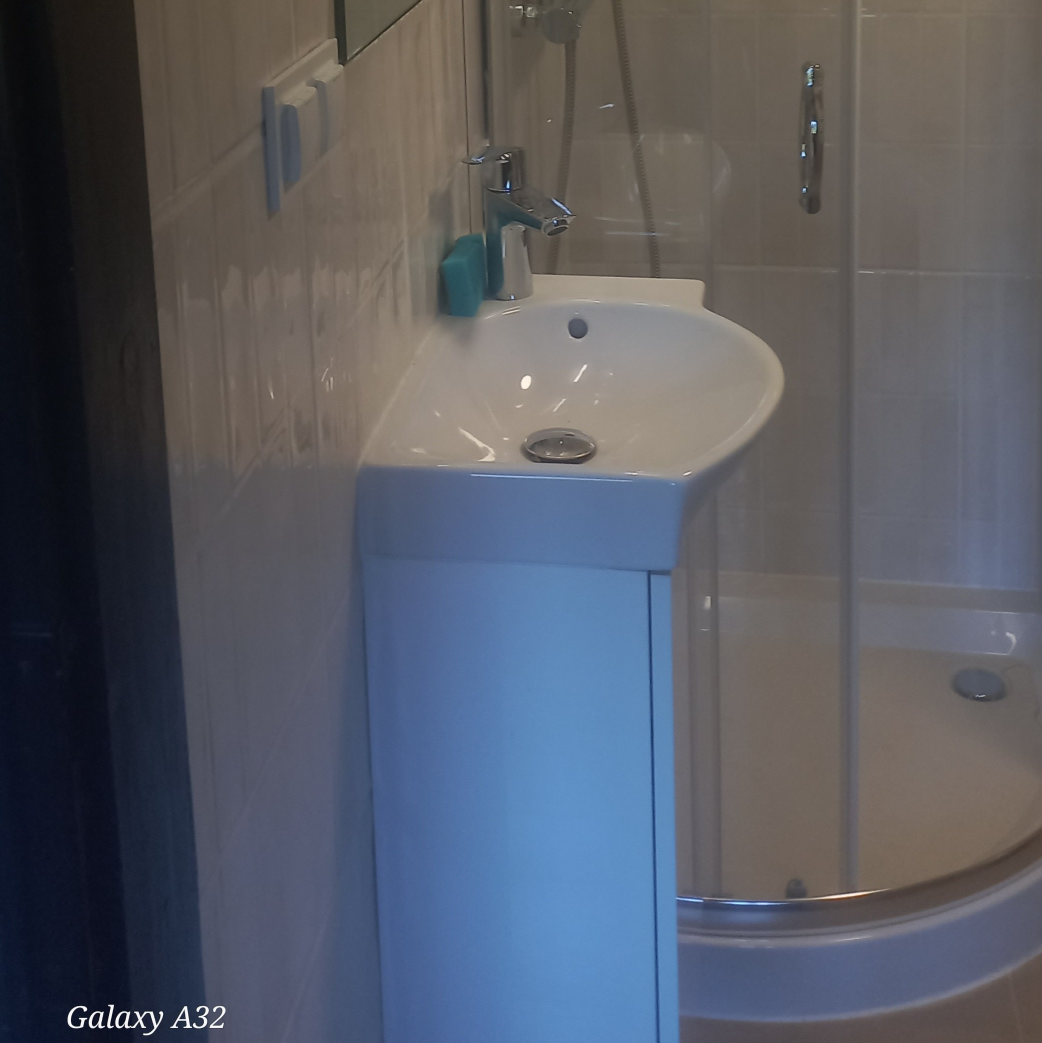 Biała umywalka z szafką w wykończonej łazience, obok kabina prysznicowa z chromowanymi elementami. Błyszczące, jasne płytki na ścianach.