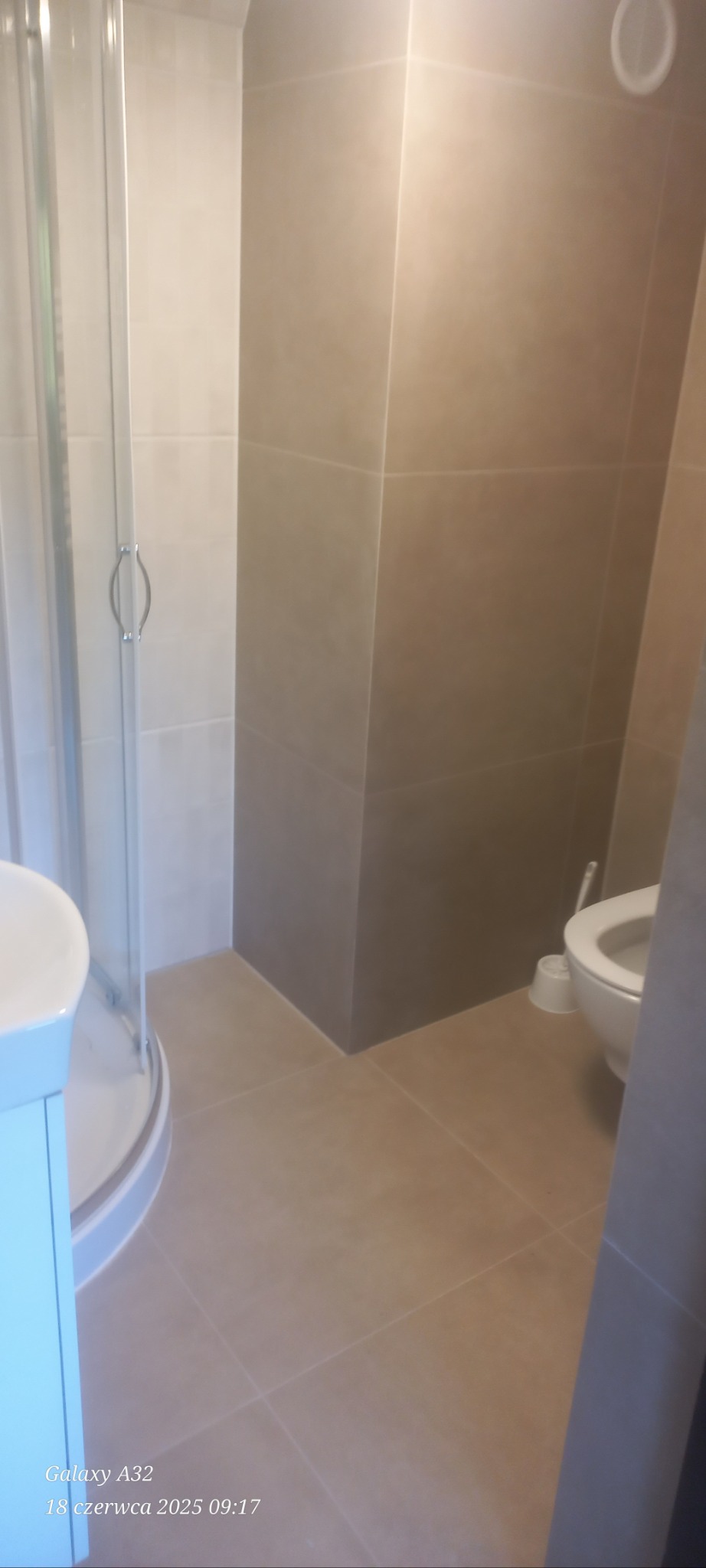 Jasna łazienka po remoncie: kabina prysznicowa z zaokrąglonymi drzwiami, beżowe płytki na ścianach i podłodze, toaleta w tle. Minimalistyczny design.