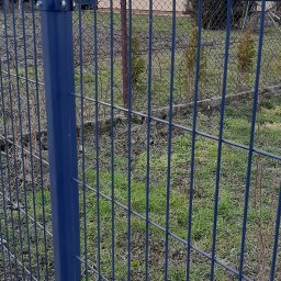 ZAKŁAD WIELOBRANŻOWY SIATKOPOL SPÓŁKA CYWILNA GRAŻYNA WILK, WOJCIECH WILK - Ciemnoszara siatka ogrodzeniowa z metalowym słupkiem na tle domu w Białobrzegach. Widoczne mocowania i fragment betonowej podmurówki.