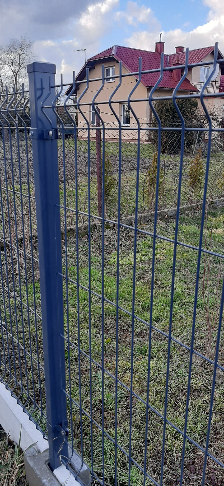 Ciemnoszara siatka ogrodzeniowa z metalowym słupkiem na tle domu w Białobrzegach. Widoczne mocowania i fragment betonowej podmurówki.