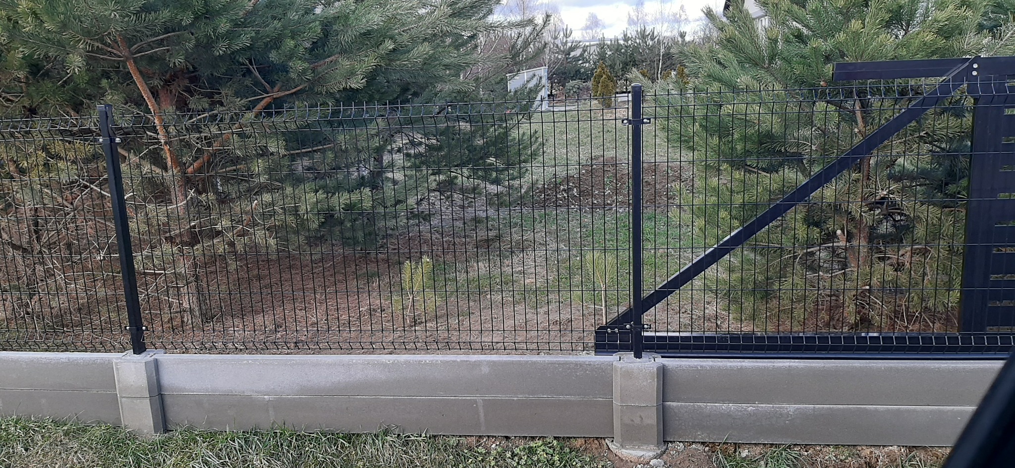 Czarna siatka ogrodzeniowa na betonowej podmurówce, z widoczną bramą, otoczona zielenią. Układ pionowy, ostry kadr, naturalne światło.