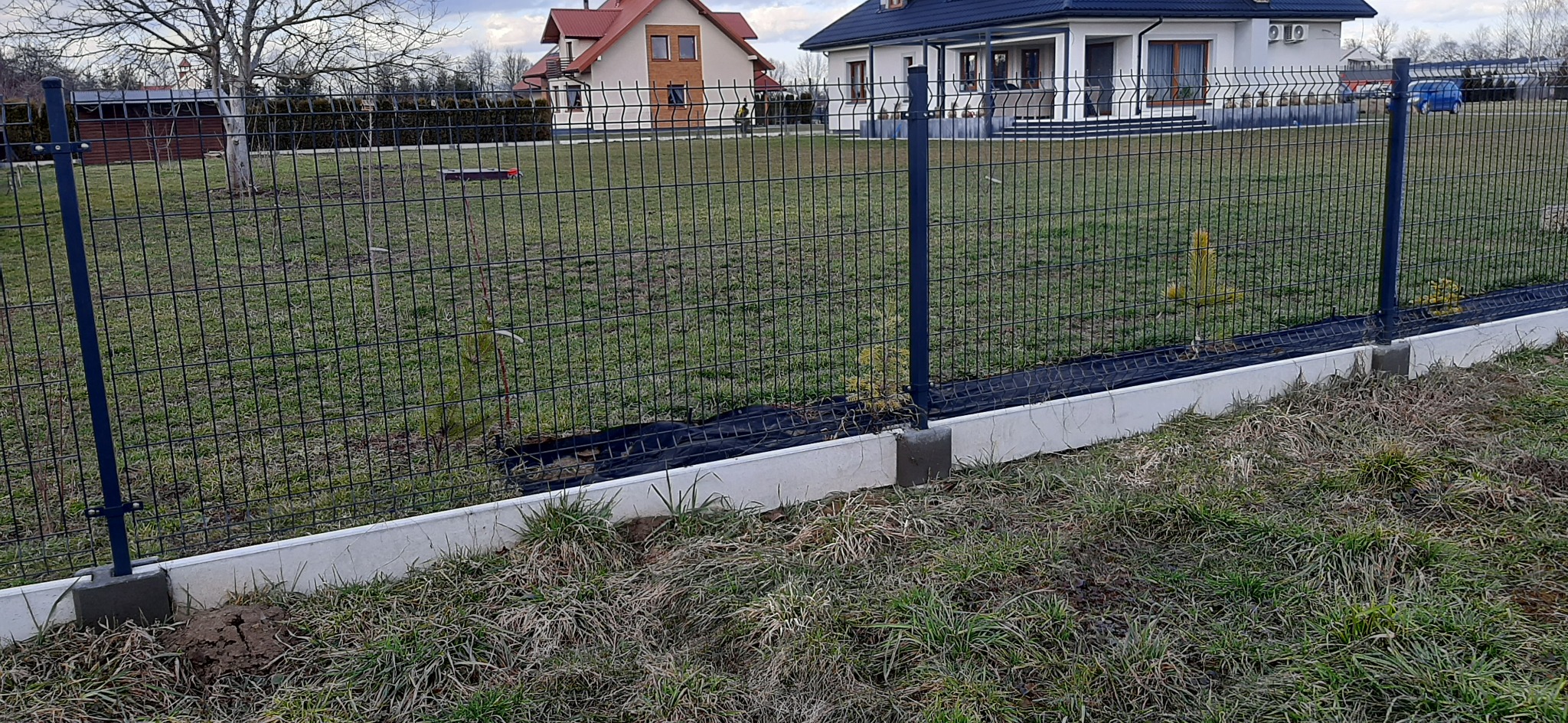Szczegółowe ujęcie ogrodzenia panelowego z podmurówką na tle domów. Widoczna siatka i słupki w kolorze grafitowym. Całość otoczona zielenią.