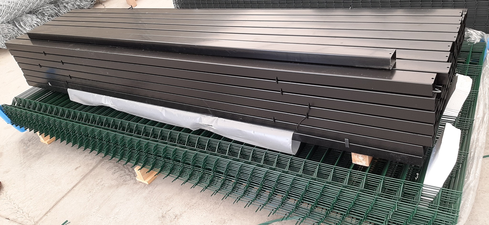 Stos czarnych, metalowych słupków ogrodzeniowych na zielonej siatce panelowej. Słupki z widocznymi otworami montażowymi, zabezpieczone folią. Elementy składowane w magazynie.