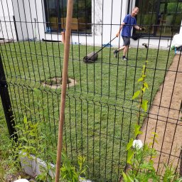 Patryk Czerniakowski - Mężczyzna w niebieskiej koszulce wyrównuje trawnik walcem ogrodowym na tle nowoczesnego domu, widok przez czarne ogrodzenie.