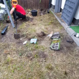 Montaż ogrodzenia: mężczyzna w czerwonej koszulce kopie dół pod słupek, obok leżą elementy ogrodzenia, narzędzia i worek z betonem B-30.