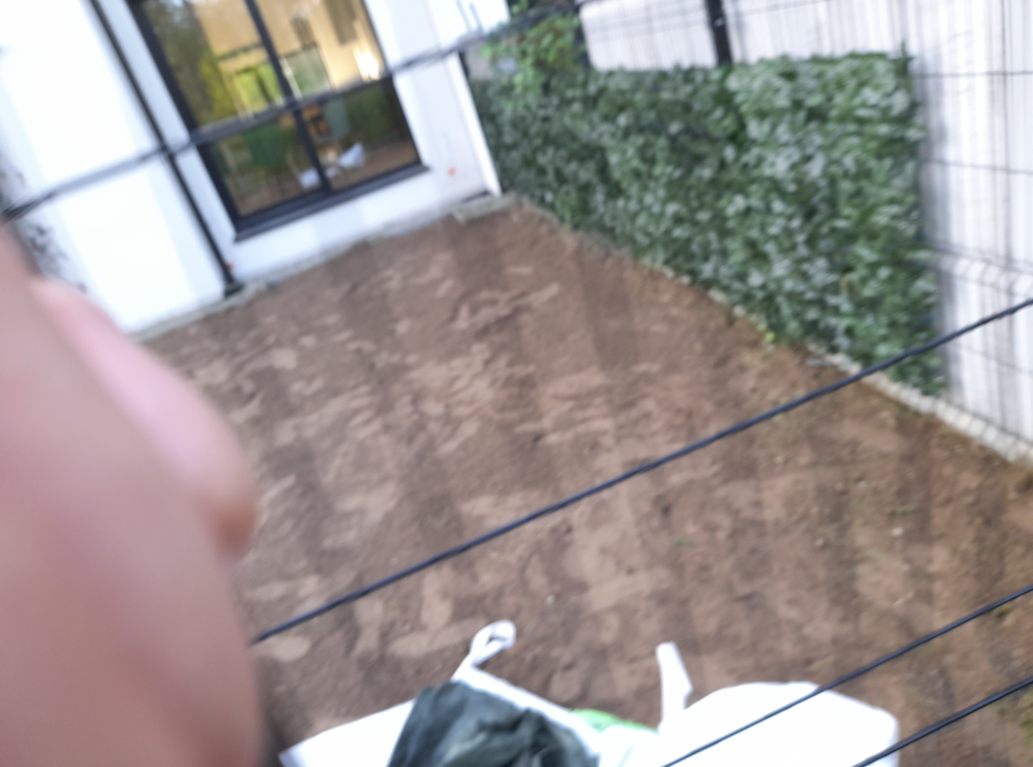 Przygotowany teren pod trawnik z widocznym systemem nawadniania, obok budynku z oknem. Częściowo widoczna sztuczna roślina na ogrodzeniu.