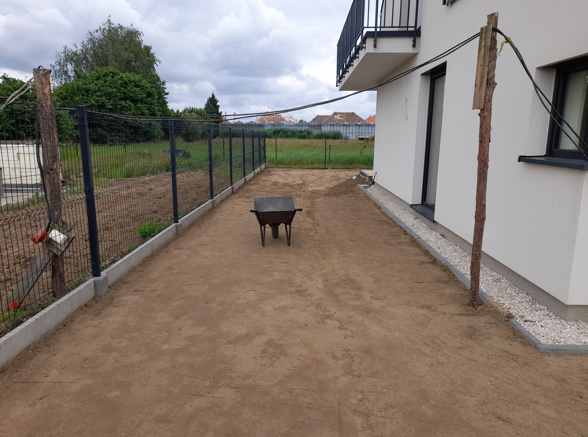 Przygotowany teren pod trawnik z czarną metalową siatką ogrodzeniową i taczką na środku, obok domu z białymi ścianami i balkonem.