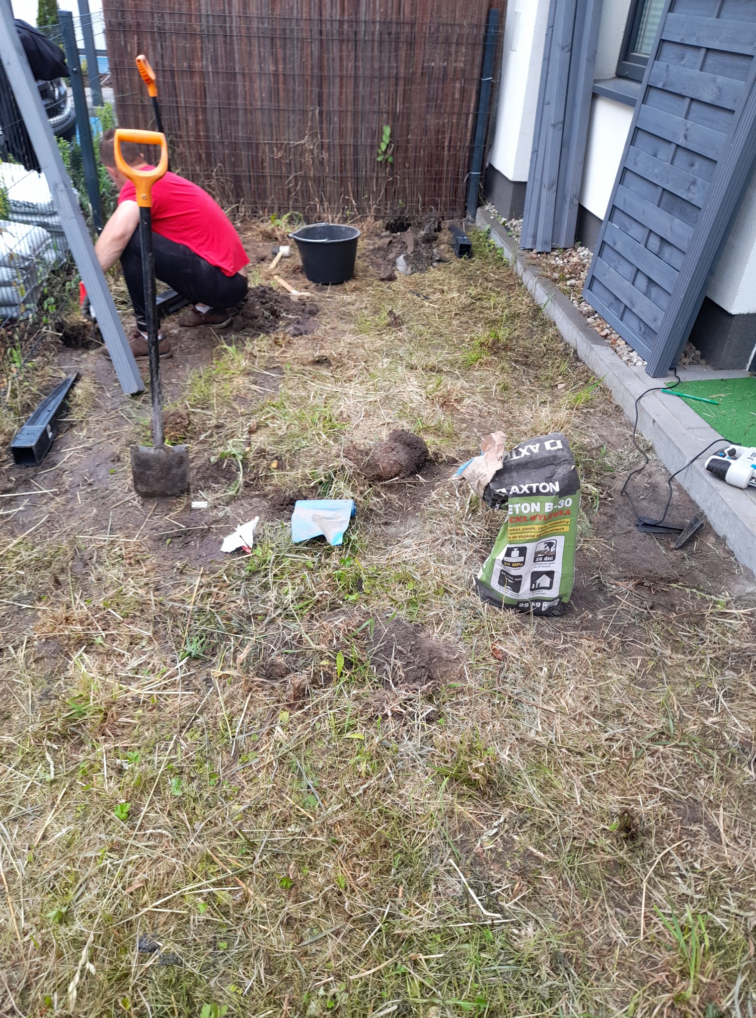 Montaż ogrodzenia: mężczyzna w czerwonej koszulce kopie dół pod słupek, obok leżą elementy ogrodzenia, narzędzia i worek z betonem B-30.
