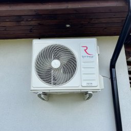 WIATRPOL SZYMON WIATROWSKI - Klimatyzator Rotenso Teta 5kW zamontowany na elewacji domu jednorodzinnego, widoczny fragment dachu z rynną. Biała ściana, błękitne niebo w tle.