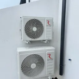 Dwa białe klimatyzatory Rotenso Teta zamontowane na białej ścianie zewnętrznej budynku. Widoczny fragment rury spustowej. Zdjęcie z bliska, w dzień.