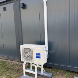 WIATRPOL SZYMON WIATROWSKI - Biała jednostka zewnętrzna klimatyzacji Haier zamontowana na metalowym stelażu przed szarą ścianą kontenera. Widoczna rura odprowadzająca.