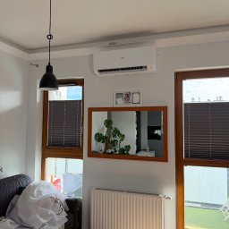 WIATRPOL SZYMON WIATROWSKI - Biała klimatyzacja Haier zamontowana nad lustrem w ramie, między oknami z brązowymi żaluzjami w jasnym salonie. Widoczny szary narożnik i biały grzejnik.