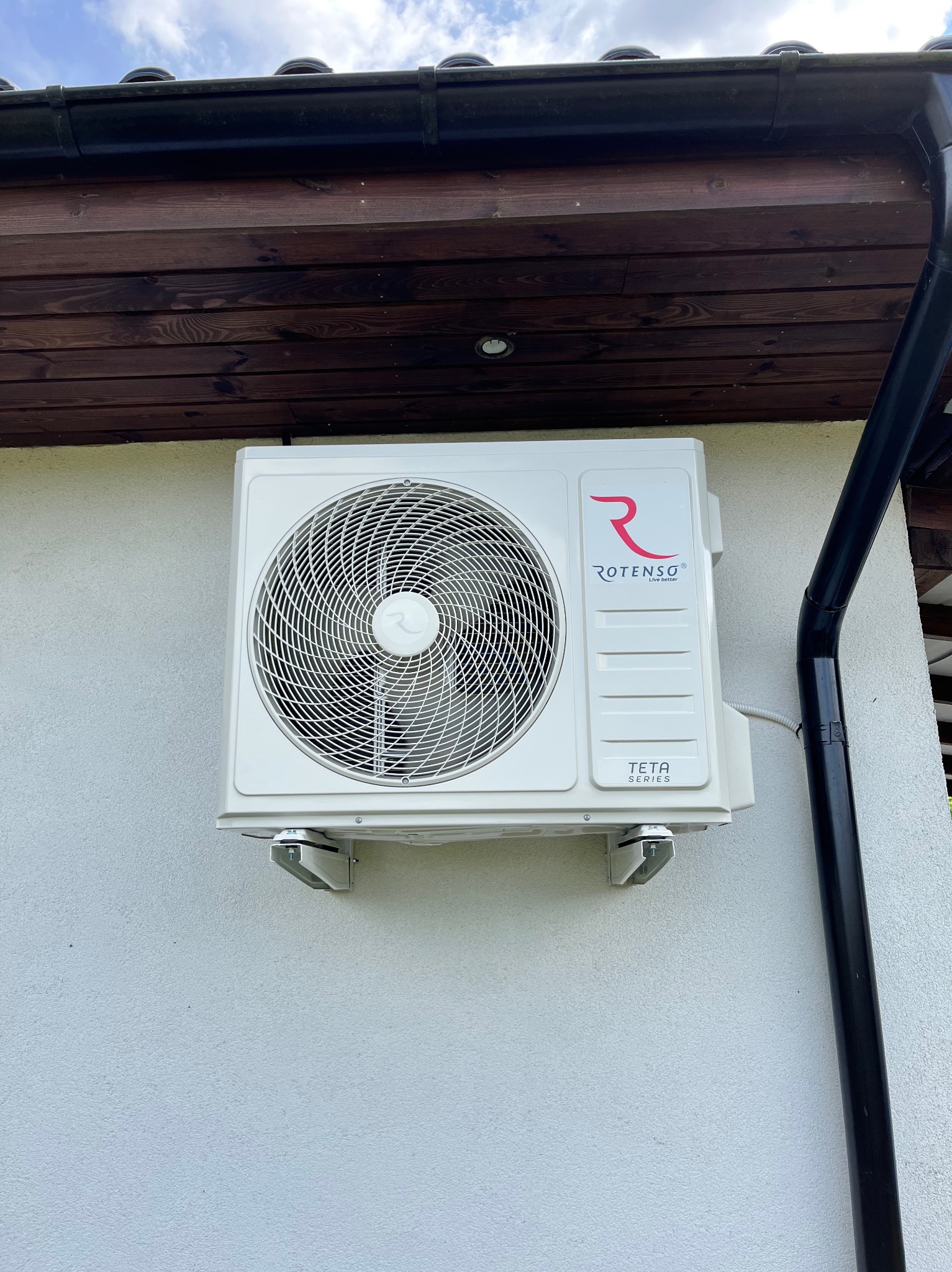 Klimatyzator Rotenso Teta 5kW zamontowany na elewacji domu jednorodzinnego, widoczny fragment dachu z rynną. Biała ściana, błękitne niebo w tle.