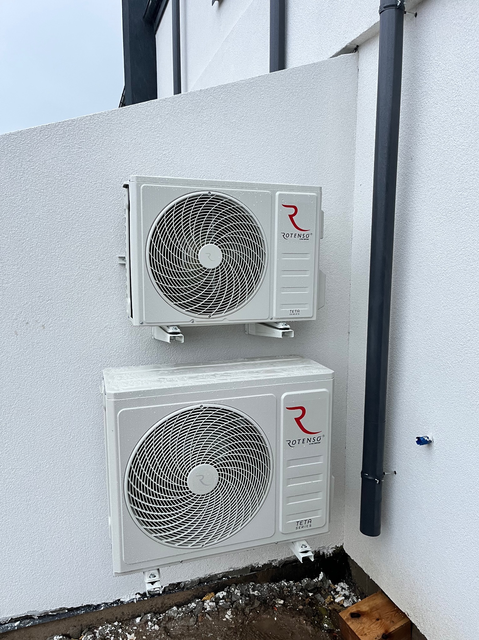 Dwa białe klimatyzatory Rotenso Teta zamontowane na białej ścianie zewnętrznej budynku. Widoczny fragment rury spustowej. Zdjęcie z bliska, w dzień.