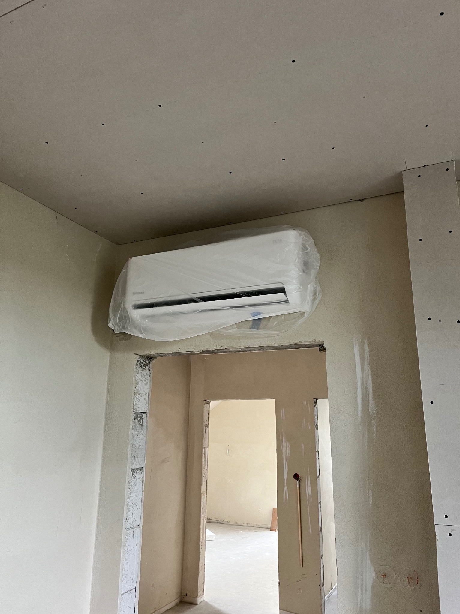 Biała klimatyzacja typu split, zabezpieczona folią ochronną, zamontowana nad wejściem w surowym wnętrzu z płyt gipsowych.