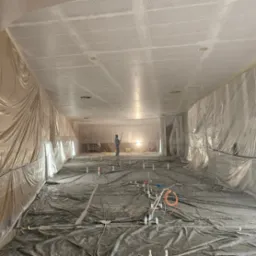 Sufit z perforacją, przygotowany do malowania natryskowego. Ściany i podłoga zabezpieczone folią. Osoba w kombinezonie maluje sufit. Widoczne instalacje podłogowe.