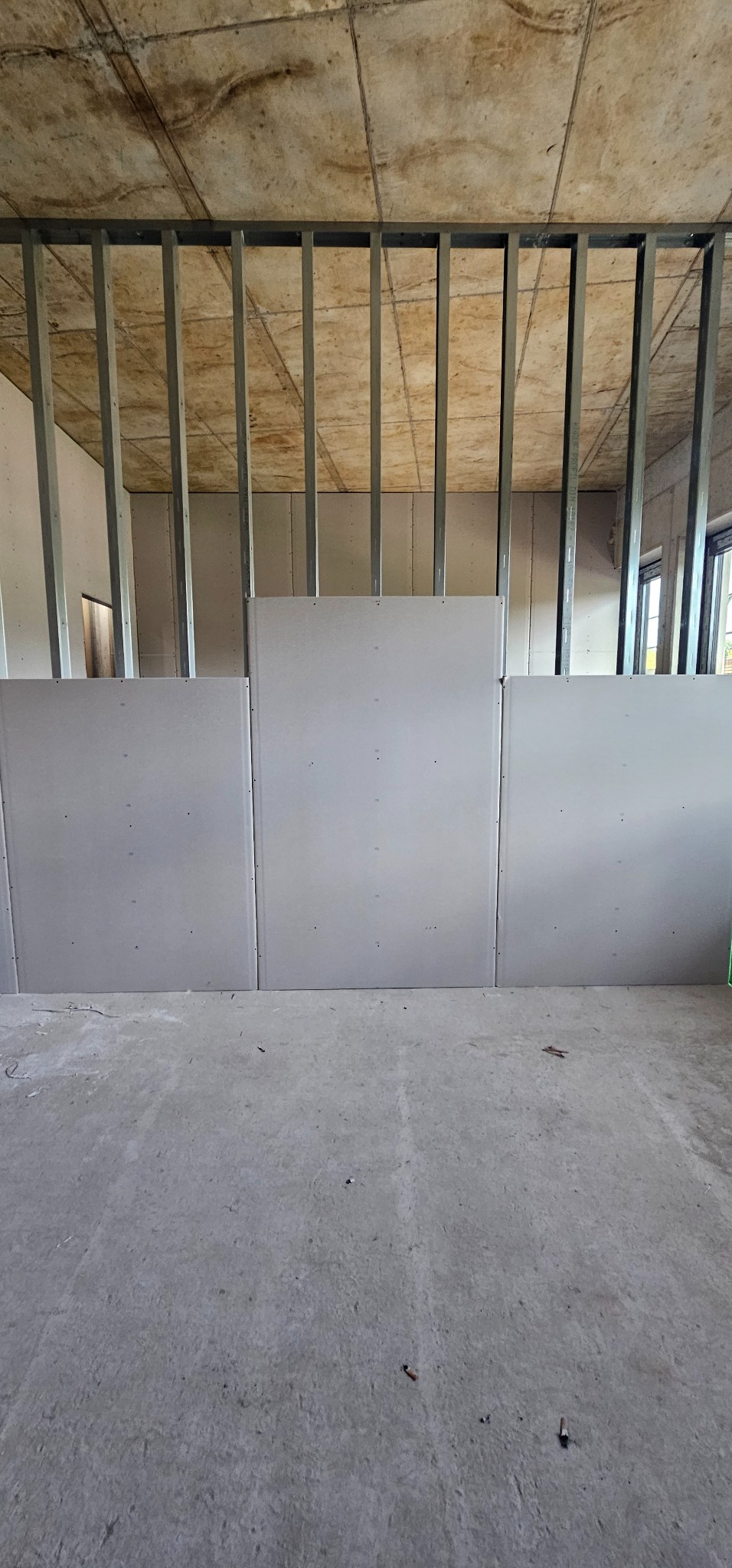 Montaż płyt karton-gips na metalowym stelażu w surowym wnętrzu. Widoczna surowa podłoga i strop. Częściowo zamontowane płyty tworzą ścianę działową.