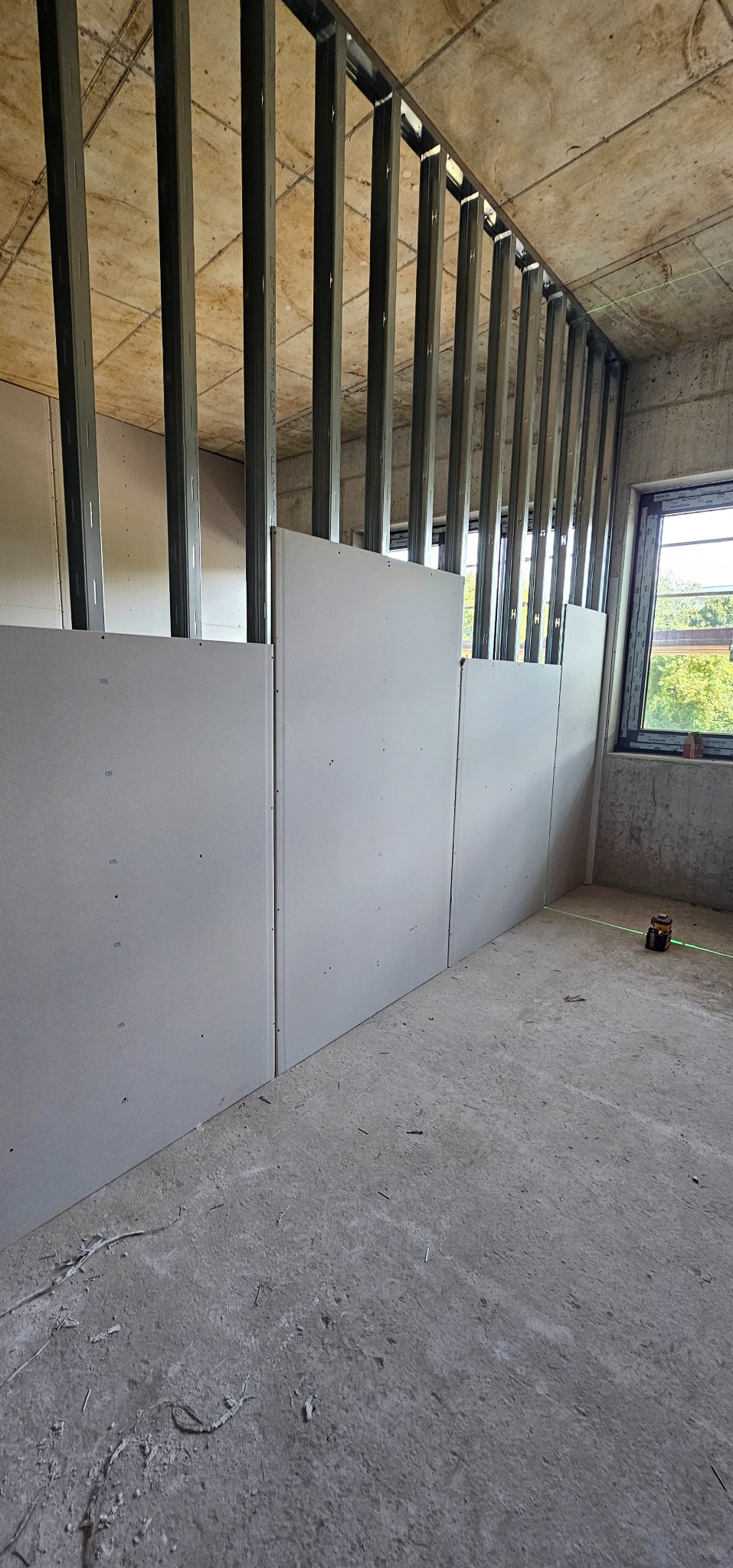 Montaż płyt gipsowo-kartonowych na metalowym ruszcie w surowym wnętrzu. Widoczne laserowe narzędzie pomiarowe, okno i surowy beton. Kątowe ujęcie.