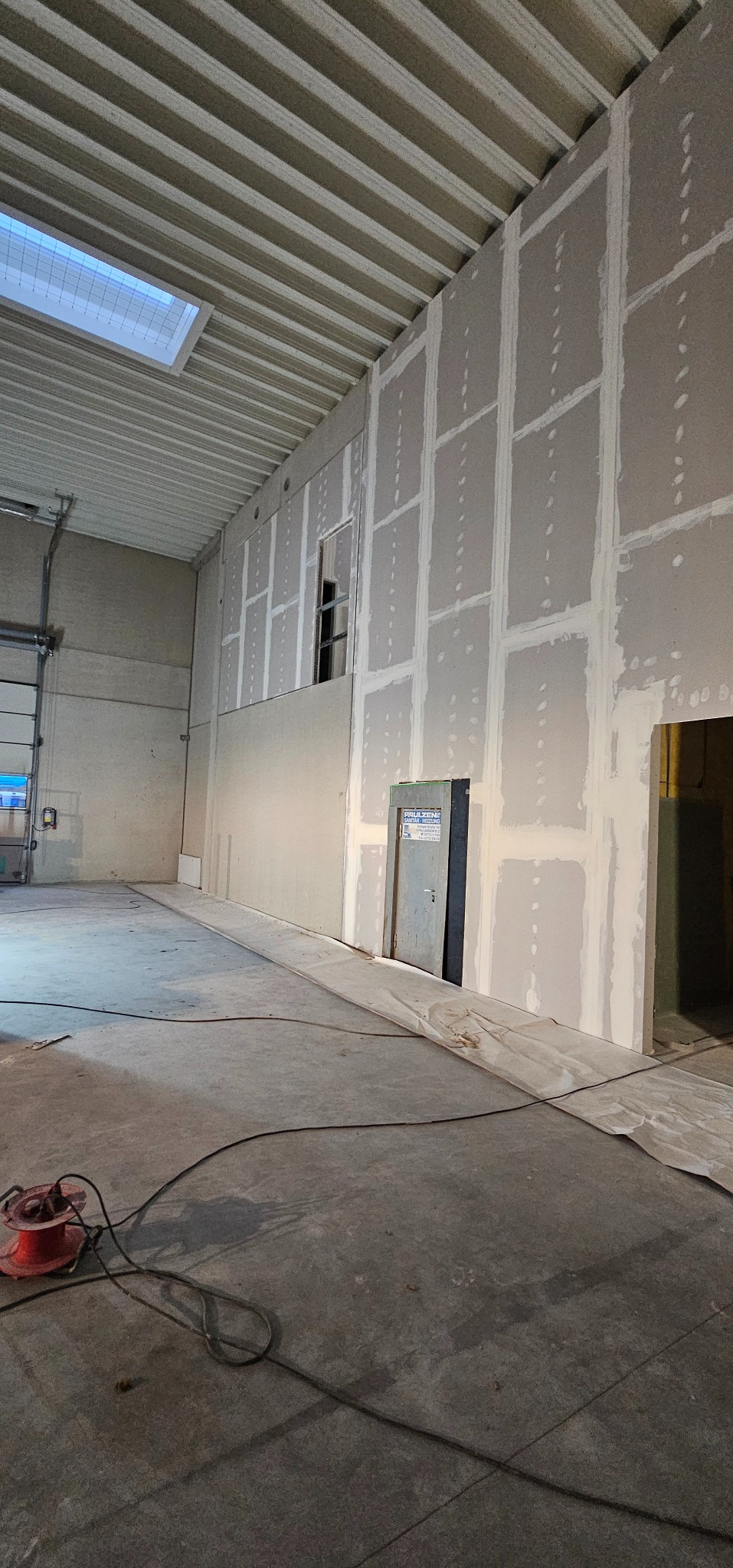 Montaż płyt karton-gips na ścianie w dużym pomieszczeniu przemysłowym. Widoczne łączenia płyt i surowy beton na podłodze. W tle brama garażowa i okno.