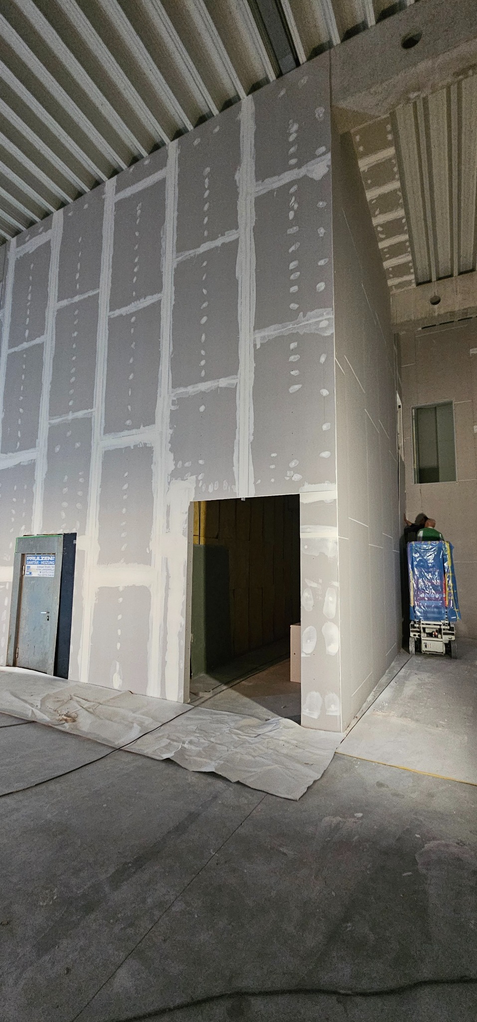 Montaż płyt gipsowo-kartonowych na ścianie z wyciętym otworem drzwiowym, widoczne łączenia szpachlowane, surowy stan wykończenia wnętrza, zabezpieczona podłoga.