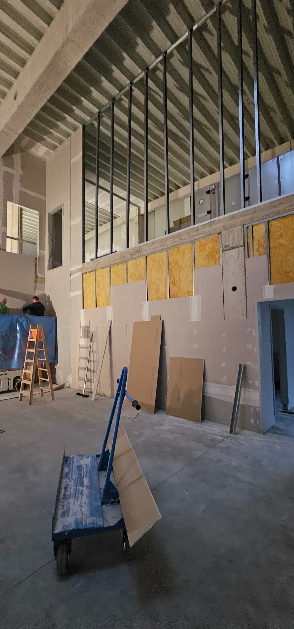 Wnętrze w trakcie prac wykończeniowych: widoczne płyty OSB, częściowo zamontowane płyty gipsowe, metalowa konstrukcja, drabina i wózek transportowy.