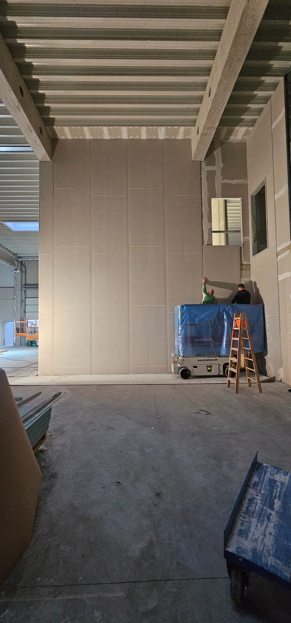 Montaż płyt karton-gips na ścianie w hali. Dwóch pracowników na podnośniku, drabina obok. Widoczne płyty, profile i surowy beton. Otwór okienny w ścianie.