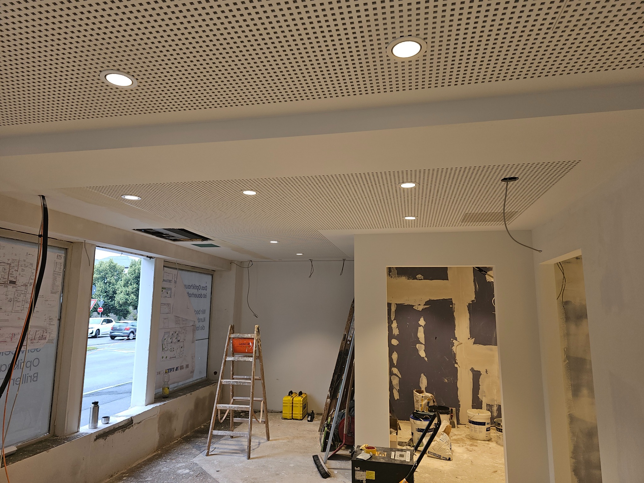 Sufit podwieszany z perforowanej płyty w trakcie montażu, widoczne oświetlenie punktowe i elementy konstrukcyjne. W tle okno i drabina.