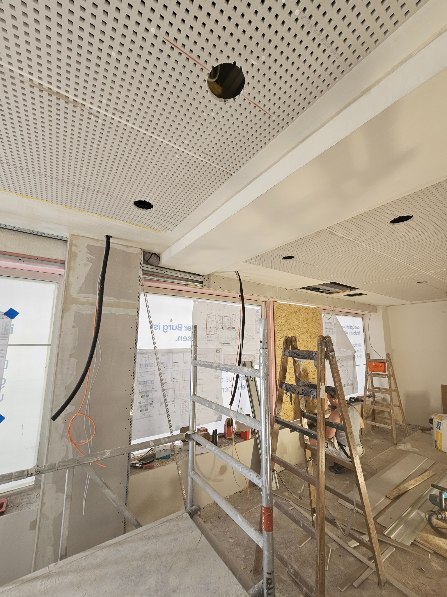 Montaż sufitu podwieszanego z perforowanej płyty w nowym biurze. Widoczne kable, rusztowania i drabiny. Prace wykończeniowe w toku.