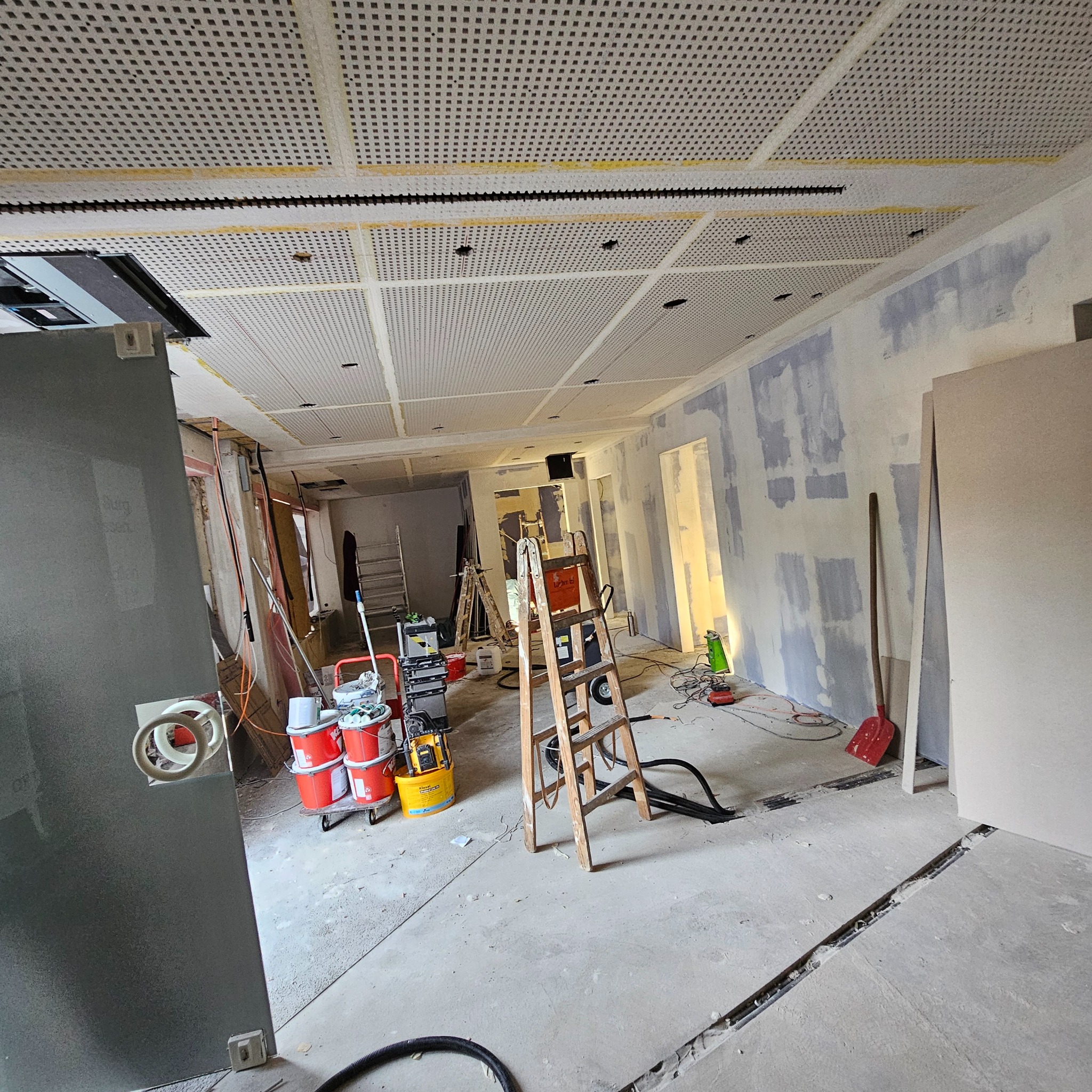 Wnętrze w trakcie remontu biura: widoczne ślady szpachlowania na ścianach, podwieszany sufit z perforacją i narzędzia budowlane, w tym drabina i wiadro z farbą.