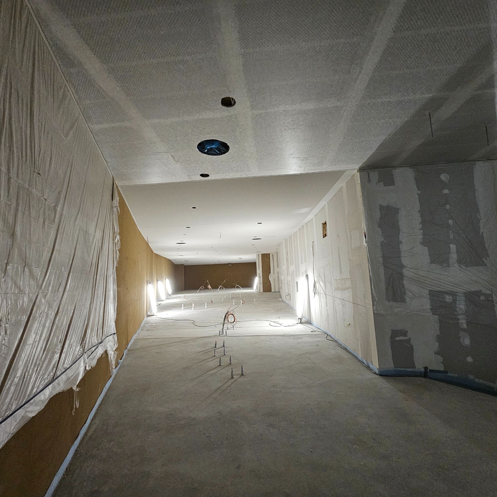 Wnętrze w trakcie prac wykończeniowych: ściany z płyt gipsowo-kartonowych, sufit z otworami na oświetlenie, widoczne instalacje podłogowe. Część ścian zabezpieczona folią.