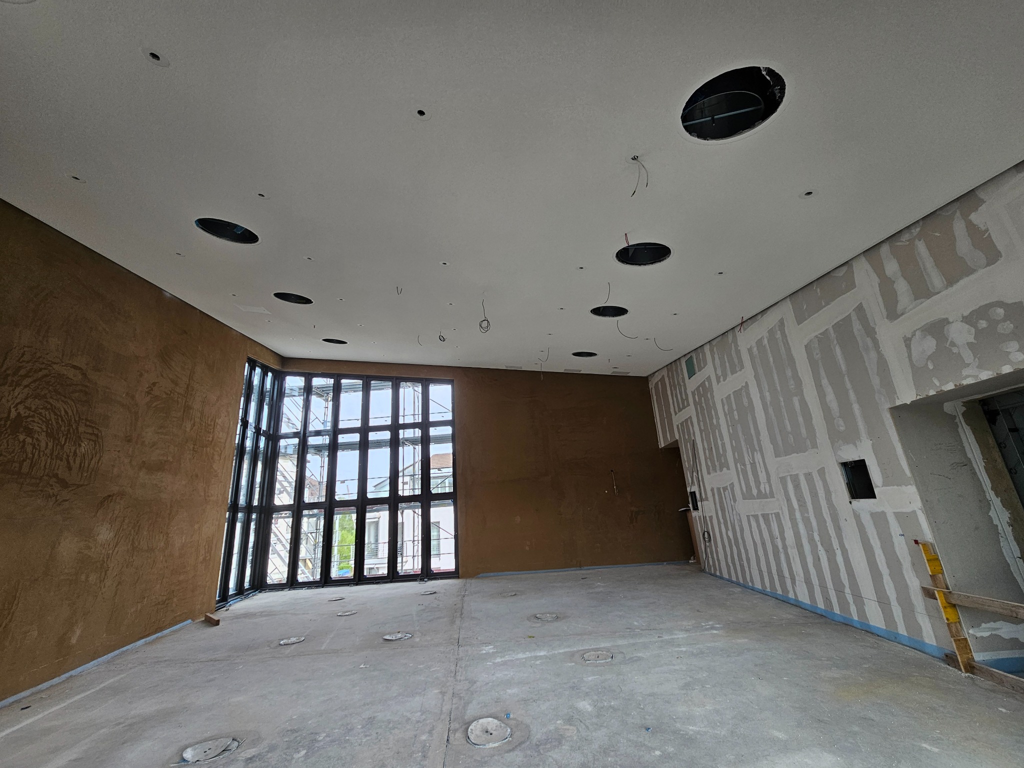 Wnętrze w trakcie prac wykończeniowych: widoczne ściany z płyt gipsowych, jedna ściana z dekoracyjną strukturą, sufit z otworami na oświetlenie i duże okno.